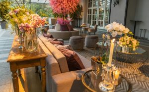 Luxe loungehoek met fluwelen poefs, bloemen in vazen en sfeervolle kaarsen in een glazen serre